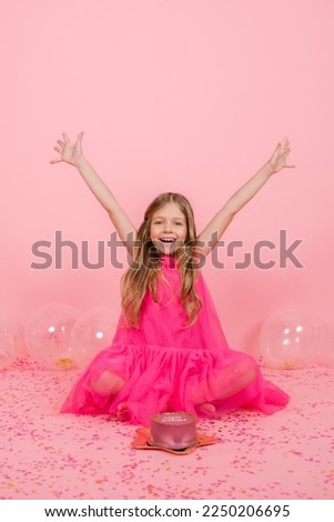 Happy smiling eight year old Caucasian girl made a wish near a bento cake for her birthday, joyfully raised her hands to the sides, sitting near balloons on a pink background with confetti