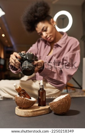 Black female photographer taking photo of bottles with oil and coconut slices on wooden board on digital camera at studio. Creating content for photostocks, commerce, social networks and advertising