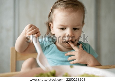 one girl small caucasian child toddler sitting at the table at home eating food alone using plastic fork childhood growing up and development concept copy space healthy eating bright photo