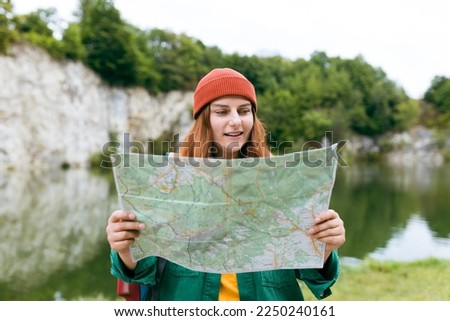 Stylish hipster woman holding paper map, wearing backpack and red hat looking at mountain view while relaxing in nature. Travel and wanderlust concept. Amazing chill moment. Full body photo