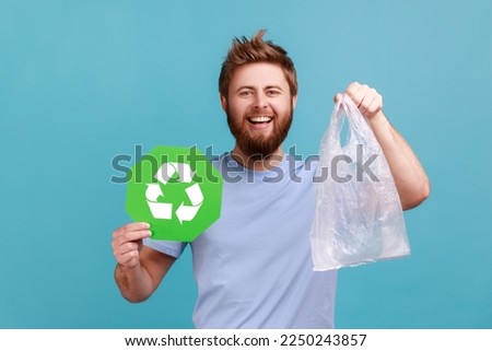 Portrait of smiling happy bearded man holding green recycling sign and plastic bag, looking at camera with positive pleasant smile. Indoor studio shot isolated on blue background.