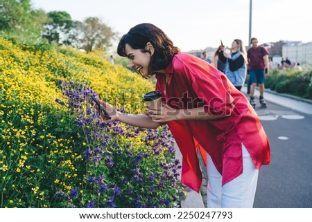 Side view of young female in casual clothes standing with cup of hot drink and pointing finger on smartphone while taking picture of blooming lavender flowers