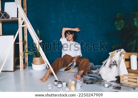 Excited young female artist sitting  on studio floor with paints enjoying her hobby.  Creative and talented brunette woman inspiring for creates picture on easel. Learning to draw with oil