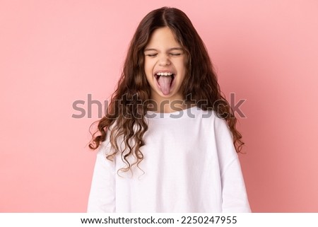 Portrait of childish carefree little girl wearing white T-shirt showing out tongue and closing eyes with naughty disobedient grimace, making face. Indoor studio shot isolated on pink background. Royalty-Free Stock Photo #2250247955