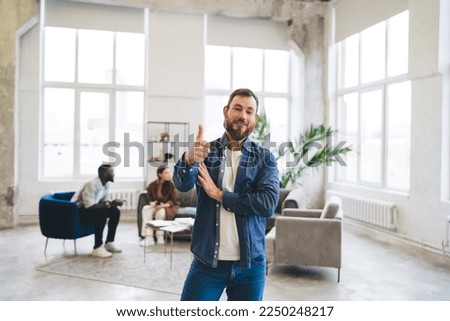 Positive bearded male employee in casual clothes showing thumb up and looking at camera while standing in modern loft workspace with coworkers in background