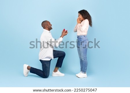 Marry me. Black middle aged man holding giving open box with engagement ring to excited young woman, asking her to be his wife during romantic date standing on one knee, blue studio background Royalty-Free Stock Photo #2250275047
