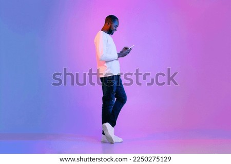 Cheerful smiling handsome middle aged black man in casual posing on neon light background, using cell phone, checking newest mobile app, surfing on Internet, full length studio shot, copy space