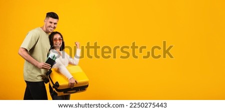 Cheerful millennial middle eastern lady in glasses with suitcase make victory, male with passports, tickets enjoy travel isolated on orange background, panorama. Sign of success, trip on vacation