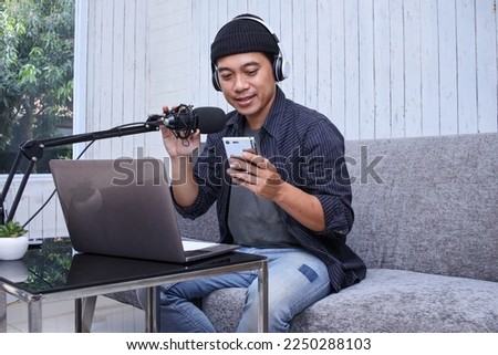 Asian man recording a podcast with headphone and microphone on his laptop while holding mobile phone. Podcaster making audio podcast from home studio.