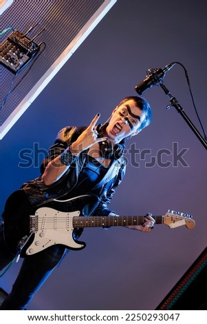 Punk rocker doing rock sign and playing guitar, feeling ecstatic and doing solo concert in studio over background. Female singer showing rock and roll symbol at live show performance.