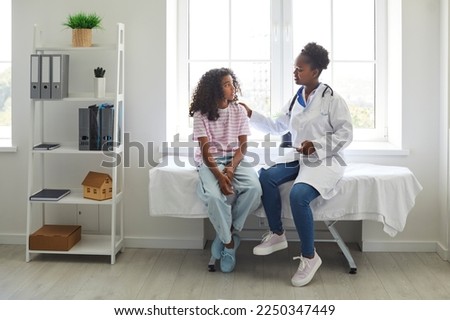 Caring, understanding doctor touches shoulder of excited teenage girl who is worried before medical examination. African American girl is sitting on examination couch and talking to female doctor. Royalty-Free Stock Photo #2250347449