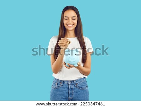 Portrait of happy woman who puts coin in piggy bank, saving money for purchase she dreams of. Young woman in casual clothes holding piggy bank in shape of pig isolated on light blue background. Royalty-Free Stock Photo #2250347461