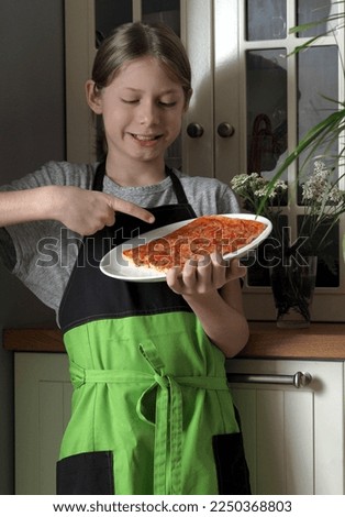 A young boy in an apron holds in his hands and demonstrates a pizza cooked at home against the background of a kitchen cabinet. Healthy home food.
