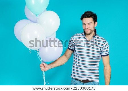 Positive Tranquil Caucasian Handsome Brunet Man With Bunch of Colorful Air Balloons Posing Against Blue Background. Horizontal Image