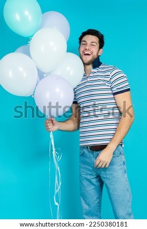 One Positive Tranquil Caucasian Handsome Brunet Man With Bunch of Colorful Air Balloons Posing Against Blue Background. Vertical Image