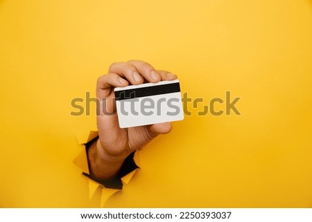Male hand is holding credit bank card in torn hole of yellow background. Online shopping, purchasing products concept.