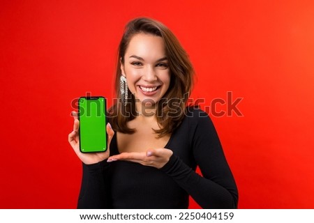 A beautiful young woman with makeup is holding a mobile phone with a green screen on a red background. Placing ads on a smartphone