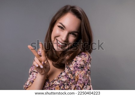 Portrait of a young woman who shows a peace sign with her fingers and smiles. A woman looks at the camera on a gray background