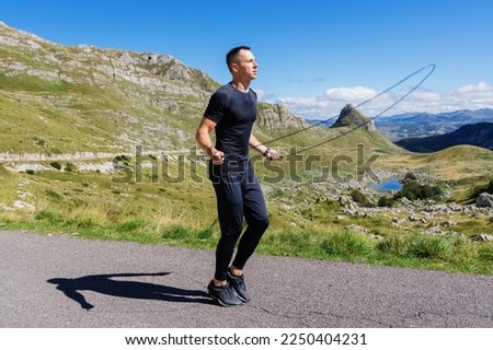An athletic man jumps on a rope against the background of mountains. Sunny day, training outside.