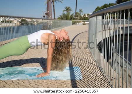 Middle-aged blonde woman, wearing green leggings and white top, doing meditation and yoga exercises on a mat outdoors. Concept yoga, meditation, concentration, sport, mindfulness.
