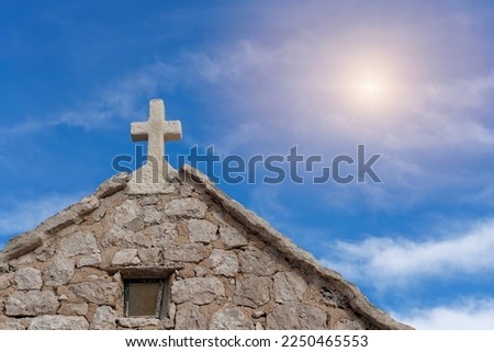 Gable of an old chapel made of natural stone with a cross on the top of the roof. Slightly cloudy blue sky backlit by the sun.