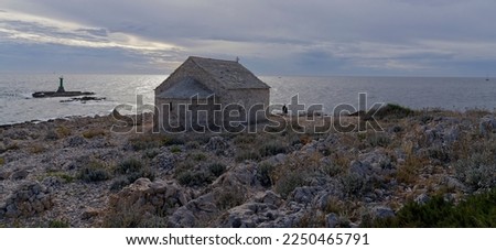 Old chapel made of natural stone on the rocky Adriatic coast of Croatia with the sea, a small lighthouse and sailboats in the background.