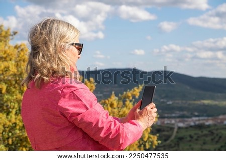 A beautiful blonde woman takes pictures of herself with her smartphone of the landscape seen from the top of a mountain that she has hiked up. Healthy life concept.