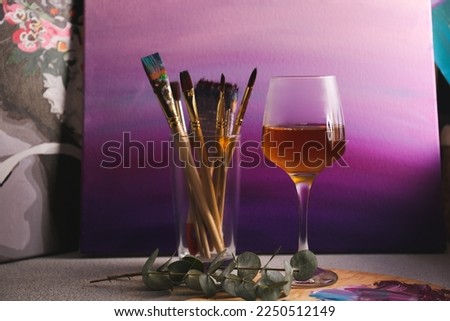Glass of tasty wine, brushes with colorful paints and gradient canvas on light gray table