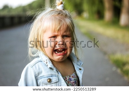A little preschool-aged 3-5 year old girl is desperately crying loudly standing on a street. The child is lost. Negative emotions. White child refugee from Ukraine. Unhappy toddler kid from orphanage. Royalty-Free Stock Photo #2250513289