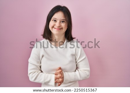Woman with down syndrome standing over pink background with hands together and crossed fingers smiling relaxed and cheerful. success and optimistic 
