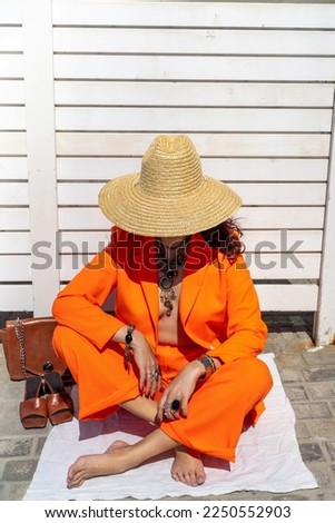 Stylish woman in an orange suit with a hat sits on a rug on a white striped background. On the hands are jewelry rings and bracelets, sandals and a bag stand side by side.