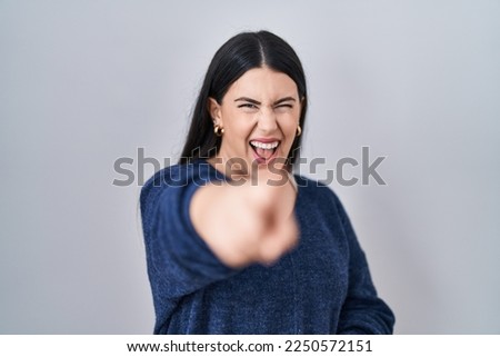 Young brunette woman standing over isolated background laughing at you, pointing finger to the camera with hand over body, shame expression 