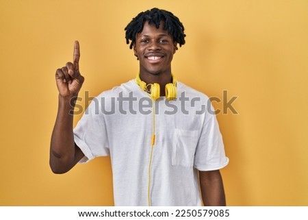 Young african man with dreadlocks standing over yellow background showing and pointing up with finger number one while smiling confident and happy. 