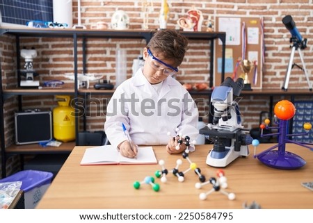 Adorable hispanic boy student writing on notebook holding molecules at laboratory classroom