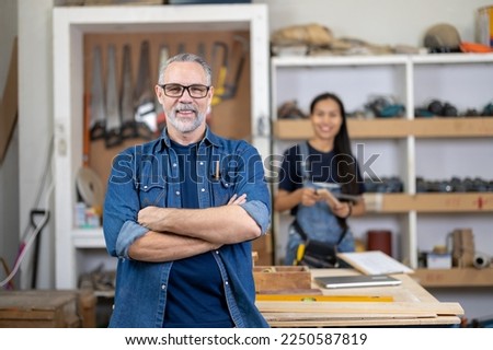 Middle ages handsome carpenter finished creating cabriolet legs and smiles to the camera with asian woman as wife stand in back, Happy Carpenter, family business owner of wood work shop.
