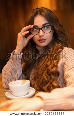 beautiful girl with glasses in a fashionable vintage knitted beige sweater sits in a cafe and drinks coffee