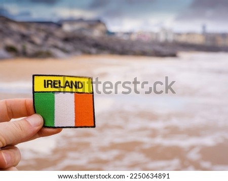 Badge with sign Ireland and Irish flag in focus. Lahinch ocean town out of focus in the background. Travel concept.
