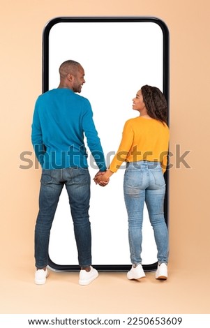 Happy black couple standing near huge phone with empty screen, standing back to camera, holding hands and smiling at each other. Great mobile app concept