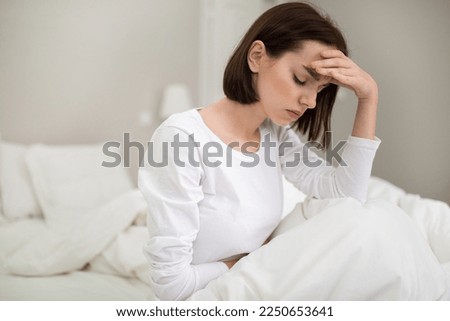 Young sick brunette woman with headache sitting in bed with closed eyes and touching her forehead at home, unhappy lady suffering from migraine or hangover, copy space Royalty-Free Stock Photo #2250653641