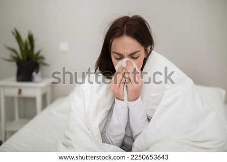 Ill unhappy millennial brunette lady in pajamas sitting in bed covered in warm blanket at home, sneezing, using napkin, suffering from pain. Cold, flu or coronavirus concept, copy space, closeup Royalty-Free Stock Photo #2250653643