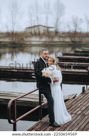 A stylish bearded groom in a black coat and a beautiful curly-haired bride in a white dress, a fur coat with a bouquet of reeds are hugging in nature, on a wooden pier. wedding photography.