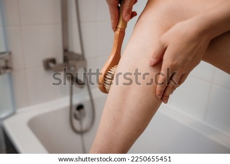 Cropped photo of a young woman in white towel doing body lymphatic drainage massage with dry wooden brush with natural bristles in bathroom at home. Anti-cellulite exercises