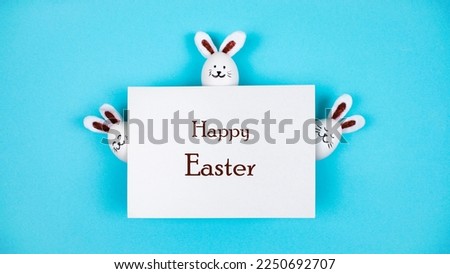 Three Easter bunnies made of white chicken eggs near sheet of white paper. Holiday greeting card "Happy Easter". Blue background. Bunny Day. Copy space.