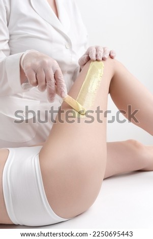 Unrecognizable beautician in white coat applying green hot wax on woman leg using spatula while woman lying down on couch. Depilation with hot wax in beauty salon. Part of photo series.