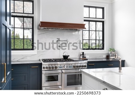 A luxurious kitchen with blue cabinets, a stainless steel stove, granite backsplash and countertop, and black framed windows. Royalty-Free Stock Photo #2250698373