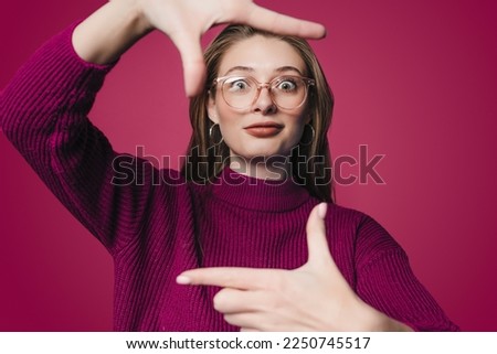 Young beautiful woman smiling, looking at camera and making frame with hands and fingers standing over crimson background. Creativity and photography concept.