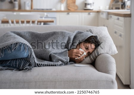 Young woman lying under blanket on sofa coughing and sneezing, using paper tissue to blow her nose. Unhealthy female caught cold or flu, resting on couch, suffering coronavirus. Cold homes and health Royalty-Free Stock Photo #2250746765