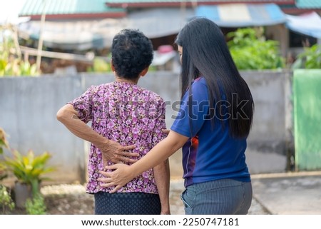 Photo of an old Asian woman standing with a young woman touching her back having back pain, rheumatism, and lumbar pain while walking in the park. Health care, health problems. Old age.