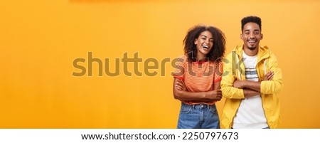 Two african american man and woman being best friends laughing out loud watching funny movie in cinema all dressed up in stylish outfit. standing with hands crossed on chest and amused expression.