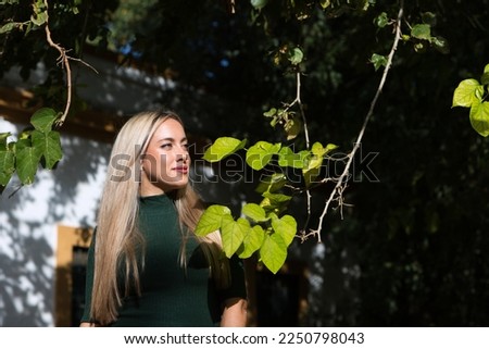 Beautiful young blonde woman dressed in a green dress is standing next to the branches of a tree through which a ray of light enters. Contact with nature.